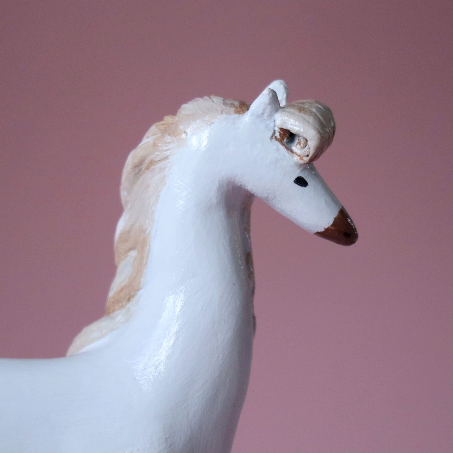 A close-up of a white horse figurine with a cream-colored mane and small brown painted details on its nose and eye area, set against a solid dusty pink background.