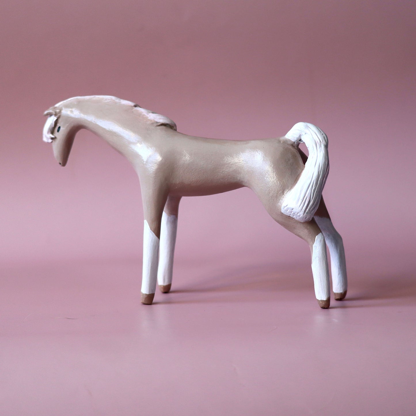A decorative horse figurine with a light beige body, white legs, mane, and tail, standing on a smooth pink studio background.