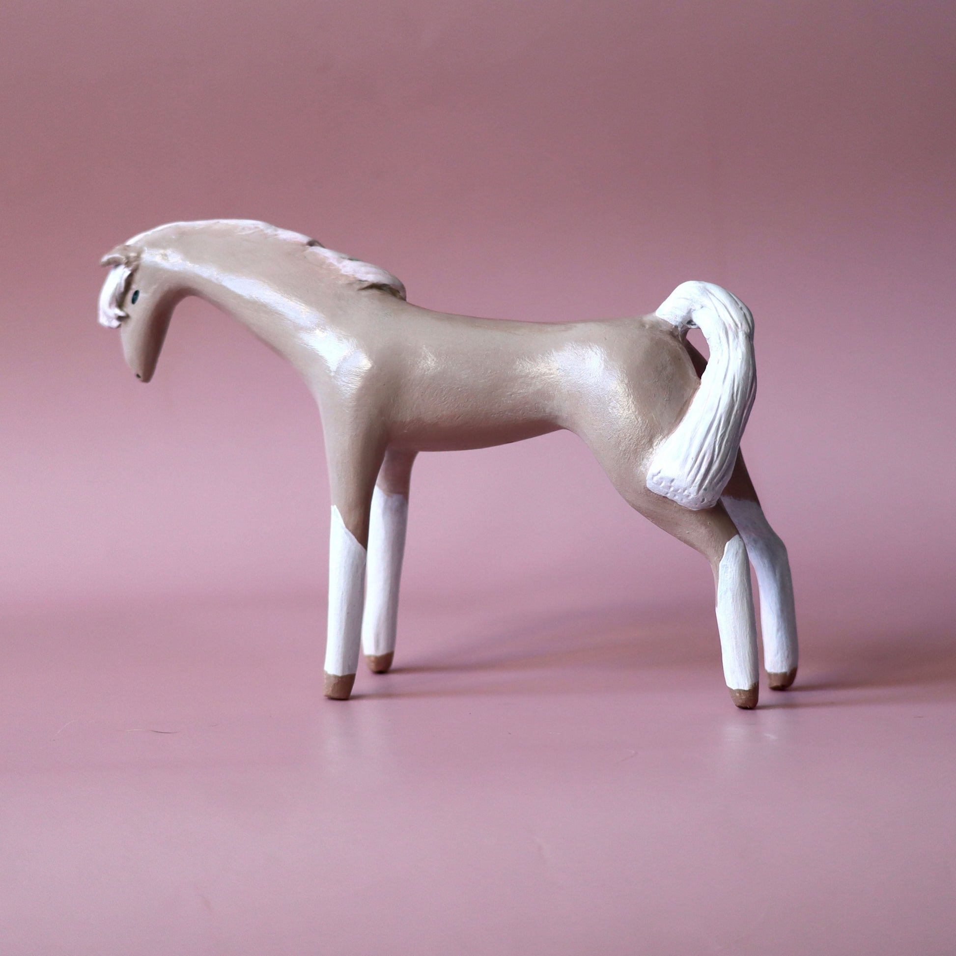 A decorative horse figurine with a light beige body, white legs, mane, and tail, standing on a smooth pink studio background.