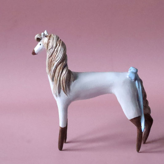 A hand-painted ceramic miniature figurine of a greyhound horse, featuring a white body, brown legs and hooves, a beige and brown mane, and a light blue tail, set against a solid dusty pink background.