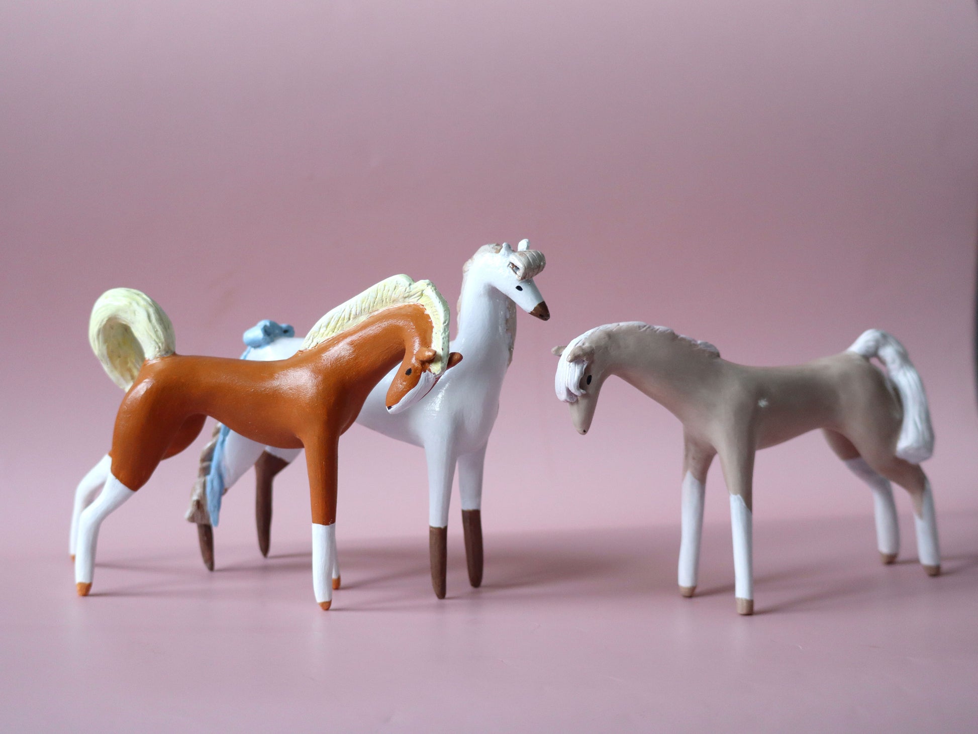 Three small horse figurines posed together on a soft pink studio background. One horse is brown with white markings and a cream mane/tail, one is white with dark brown legs, and one is light gray with a white mane and tail.