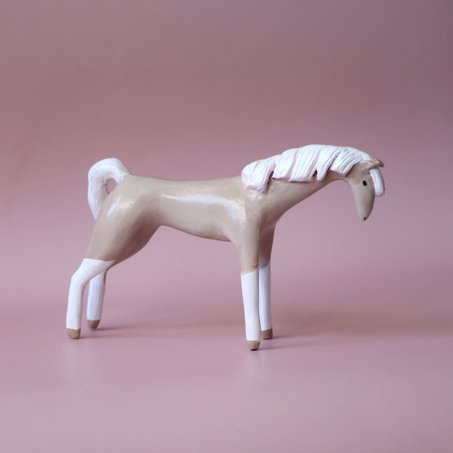 A stylized horse figurine in a side profile, featuring a beige body, white mane and legs, and small brown hooves, set against a solid dusty pink background.