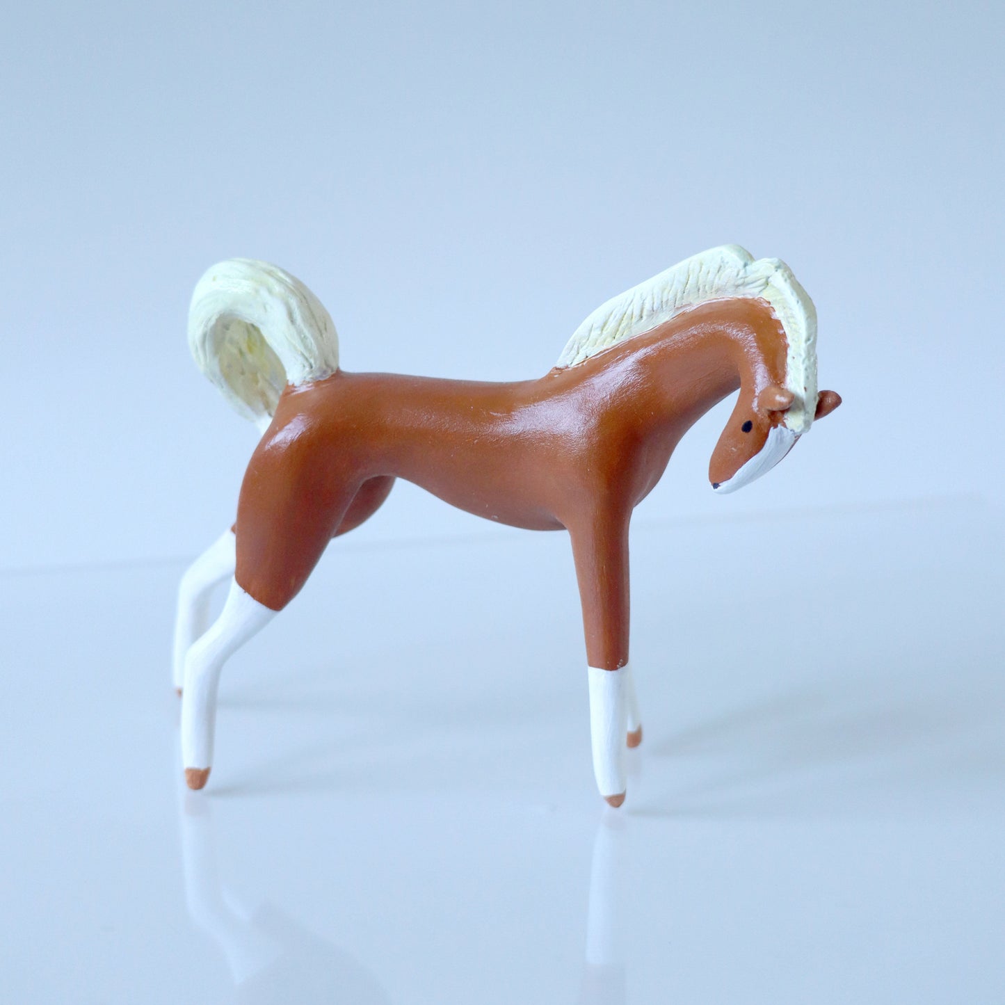 A horse sculpture in profile on a clean light gray studio background. The horse is primarily medium brown with a white mane, tail, lower legs, and face.